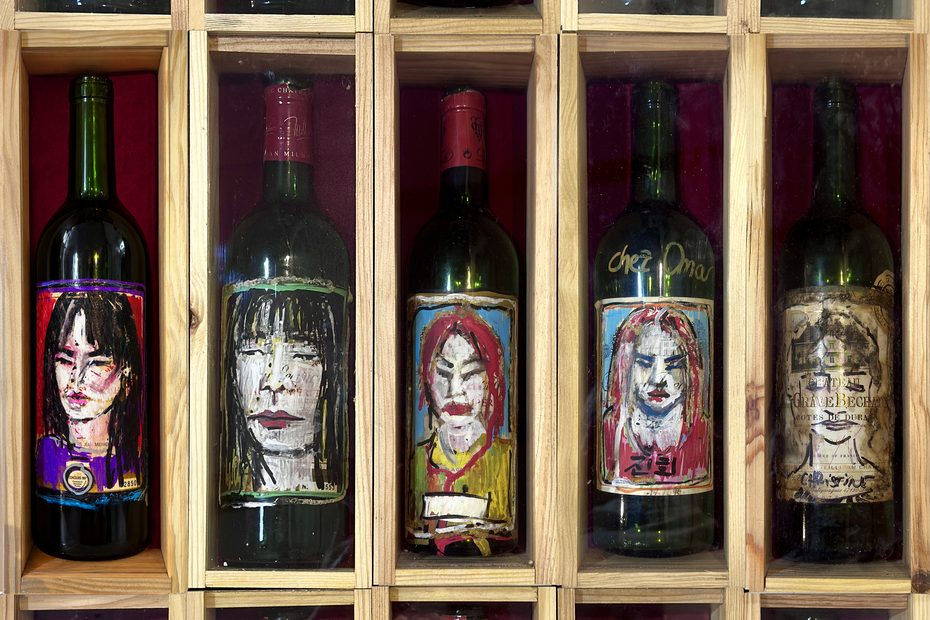 Fifteen wine bottles with hand-painted portraits on their labels, displayed in wooden boxes in three rows of five.